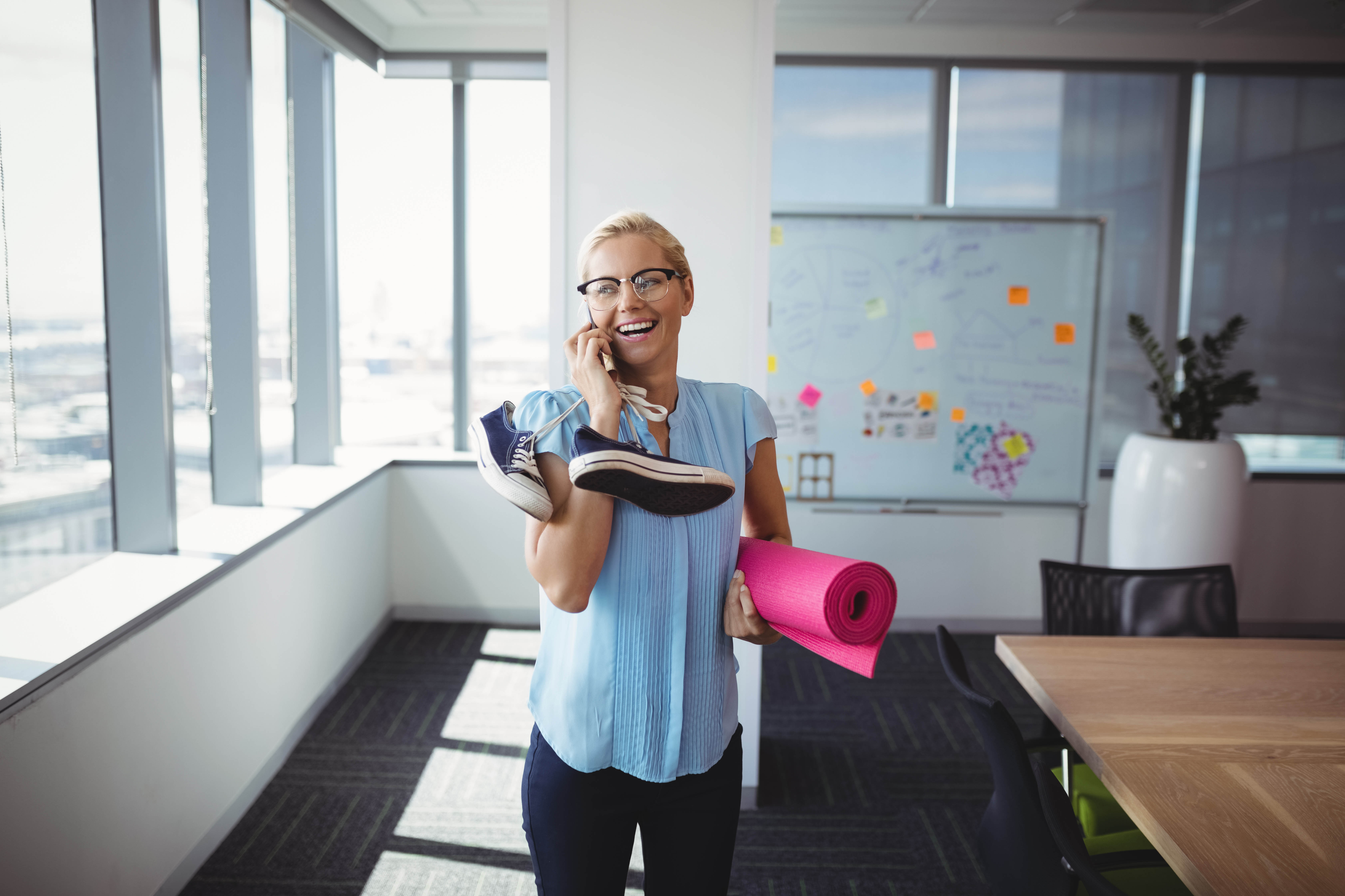 Young blond woman talks on the phone while carrying her yoga mat and running shoes in the office Young blond woman talks on the phone while carrying her yoga mat and running shoes in the office